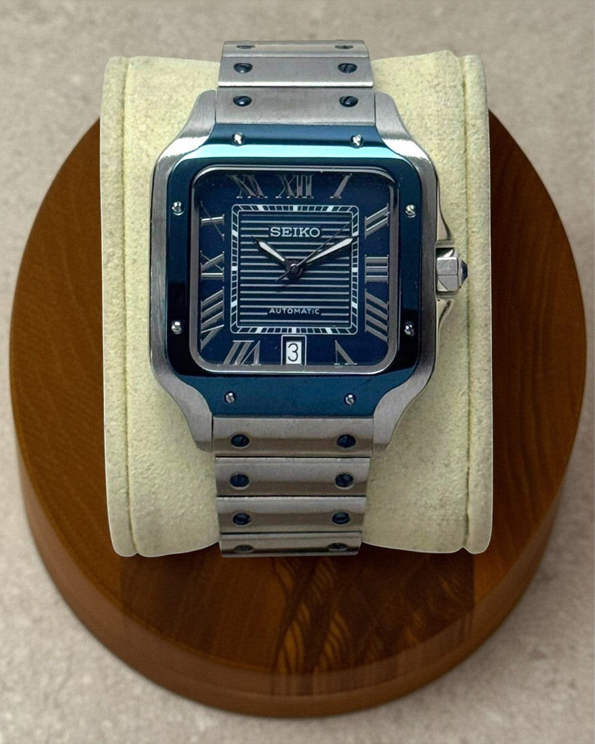 PB TimePiece Aviator watch with a navy blue dial, Roman numerals, and a stainless steel bracelet on a display stand.