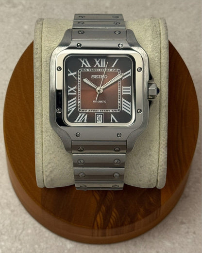 PB TimePiece Aviator watch with ombre brown dial, Roman numerals, and stainless steel bracelet on a display stand.